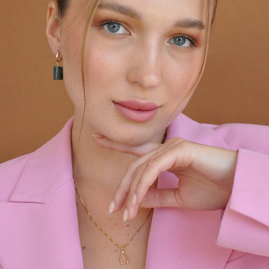 Close-up portrait of woman wearing Lykke Lucky Wishbone 14k gold filled necklace with hand near cheek in pink blazer