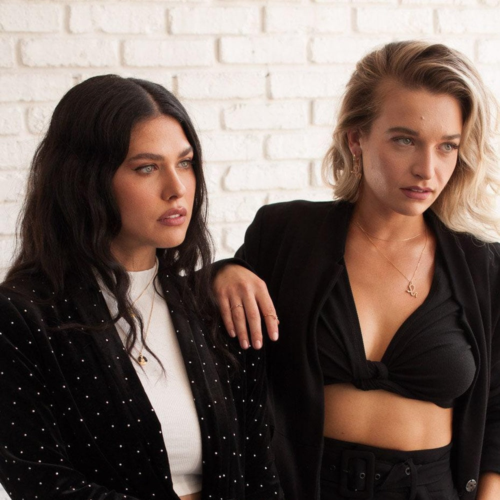 Two women posing in front of a white brick wall. The woman on the left has long dark hair and wears a black velvet blazer with silver studs over a white top.