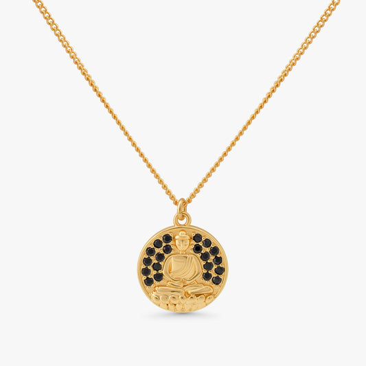 A gold pendant necklace featuring a circular medallion with a seated Buddha figure, accented by small black stones around the upper half, suspended from a delicate gold chain against a light gray background.