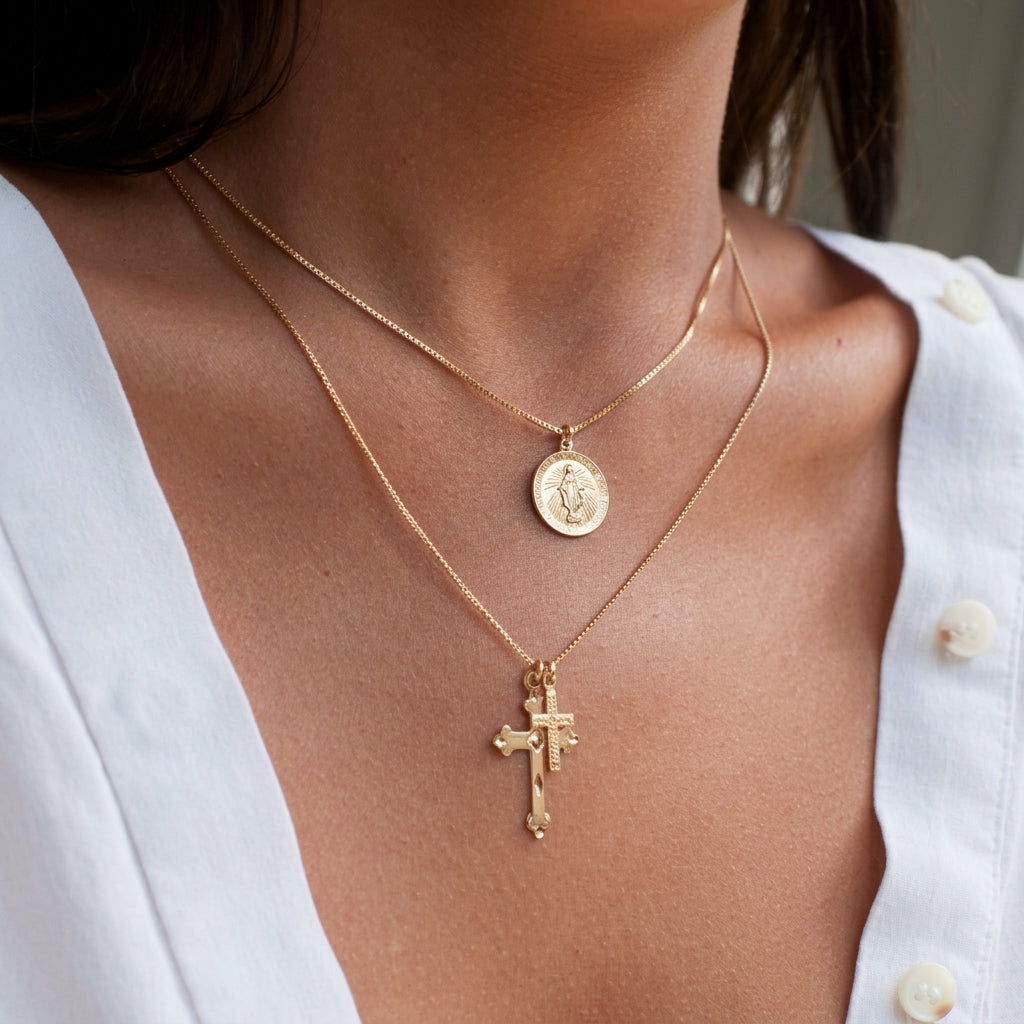 A close-up of a person wearing two layered gold necklaces: a fine chain with a round medallion pendant and a longer chain with a cross pendant, styled with a white button-up shirt.