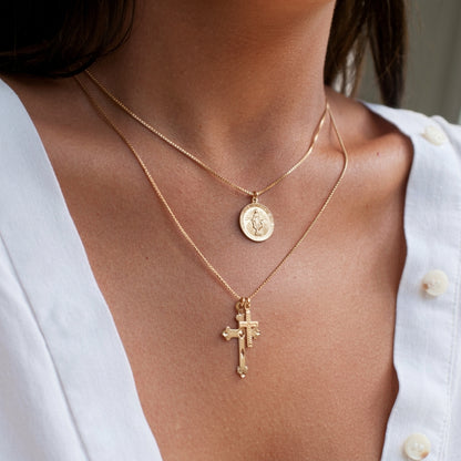 A close-up of a person wearing two layered gold necklaces: a fine chain with a round medallion pendant and a longer chain with a cross pendant, styled with a white button-up shirt.