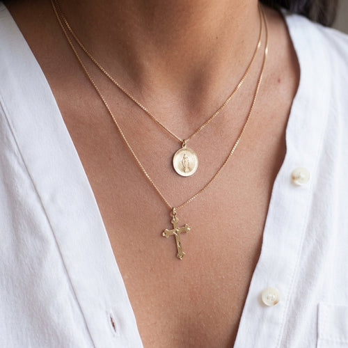 A close-up of a person's neck and chest wearing two layered gold necklaces: a short chain with a round medallion pendant and a longer chain with a cross pendant, styled with a white button-down shirt.