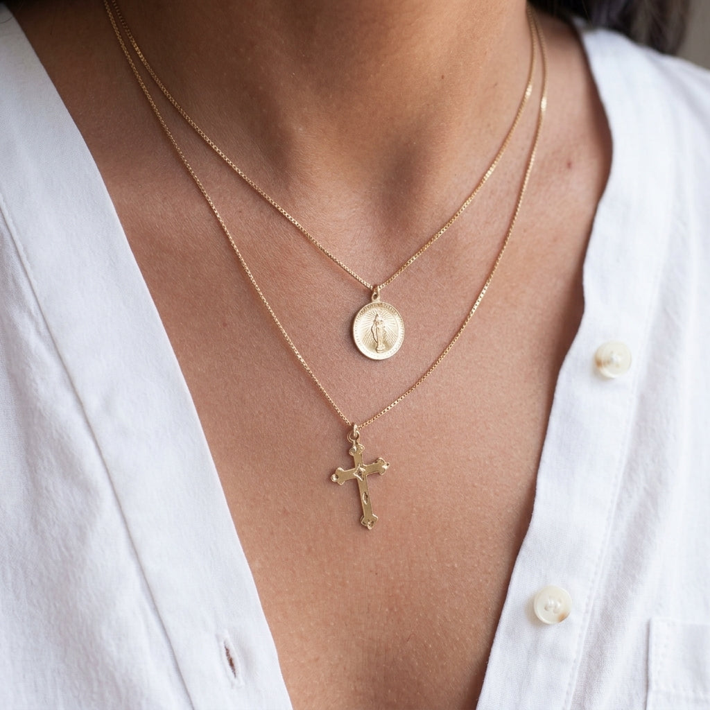 A close-up of a person's neck and chest wearing two layered gold necklaces: a short chain with a round medallion pendant and a longer chain with a cross pendant, styled with a white button-down shirt.