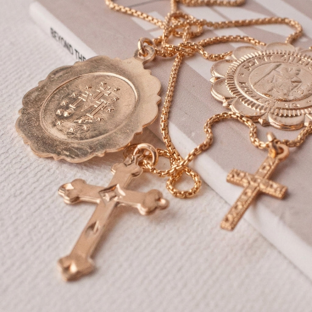 A gold-toned rosary-style necklace with a crucifix pendant, a large oval medallion pendant, and a smaller round medallion pendant, displayed on a light textured surface.
