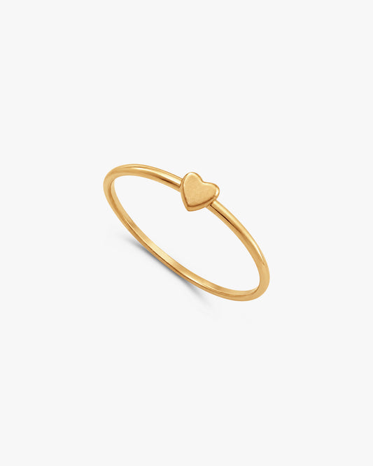 A delicate 14k gold-filled ring with a small, polished heart charm, resting on a textured white surface.