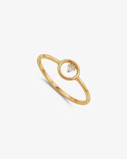 A delicate rose gold ring featuring a minimalist open circular design with a small, clear heart-shaped gemstone in the center, resting on a light textured white surface.