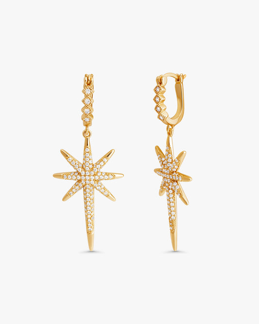 A pair of gold huggie hoop earrings with dangling starburst charms paved with small clear stones, shown on a light gray background.