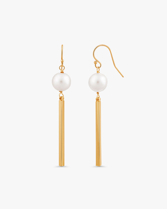 A pair of gold and white drop earrings on a light gray background. Each earring features a white pearl suspended from a polished gold fishhook ear wire and a slim, cylindrical gold bar.