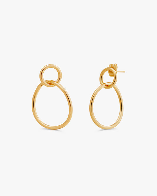 A pair of gold-toned drop earrings with a large oval and a smaller interlocking ring, shown on a clean off-white background.