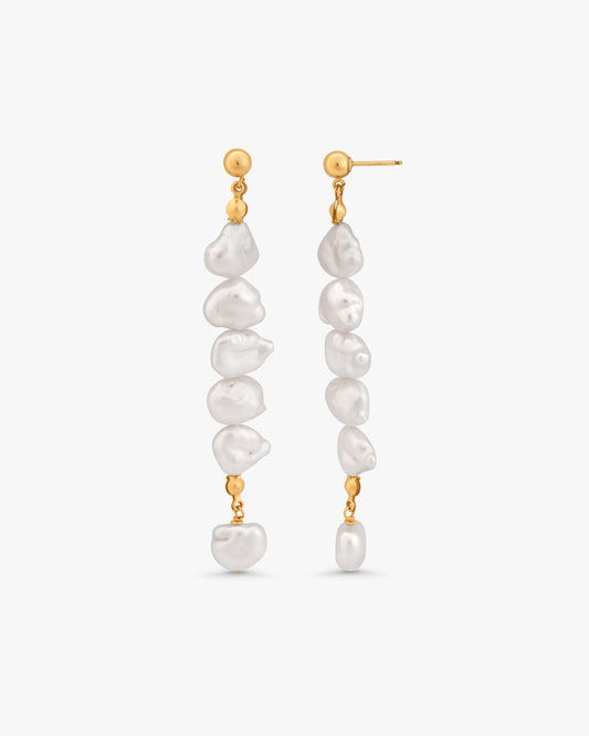 A pair of gold dangle earrings with irregular white baroque pearls, shown against a light gray background.