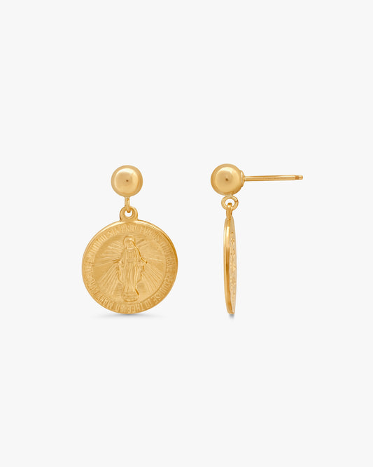 A pair of gold earrings on a light gray background, showing one front-facing drop earring with a circular religious medallion and one side-profile post earring.