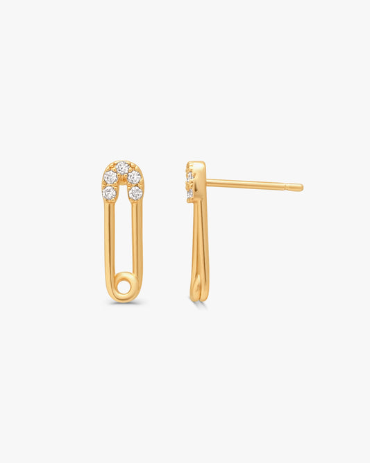 A pair of gold safety-pin stud earrings with a small cluster of clear stones at the top, shown on a light gray background.