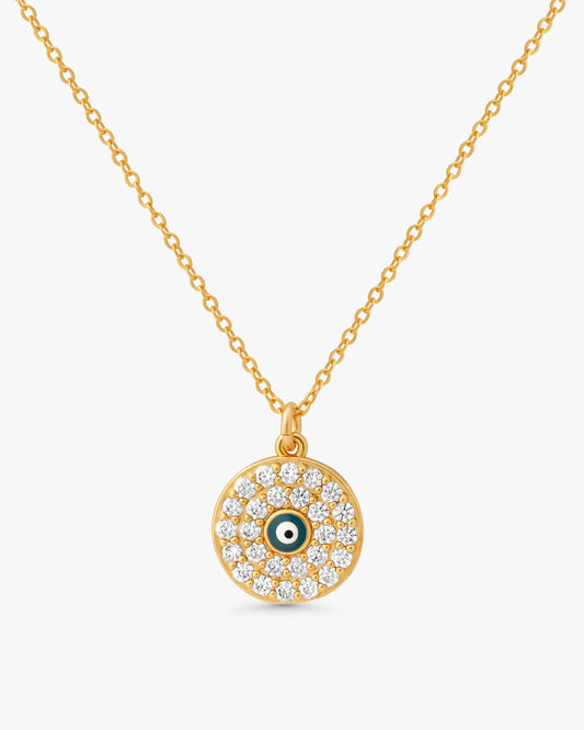 A gold chain necklace with a circular evil eye pendant, featuring clear pavé stones and a small green and white eye detail in the center, set against a light gray background.