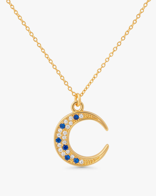 A gold crescent moon pendant necklace on a delicate gold chain, featuring small blue and clear gemstones along the curve, set against a light gray background.