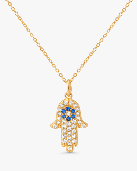 A gold Hamsa hand pendant necklace adorned with clear crystals and blue gemstones, suspended from a delicate gold chain against a light gray background.