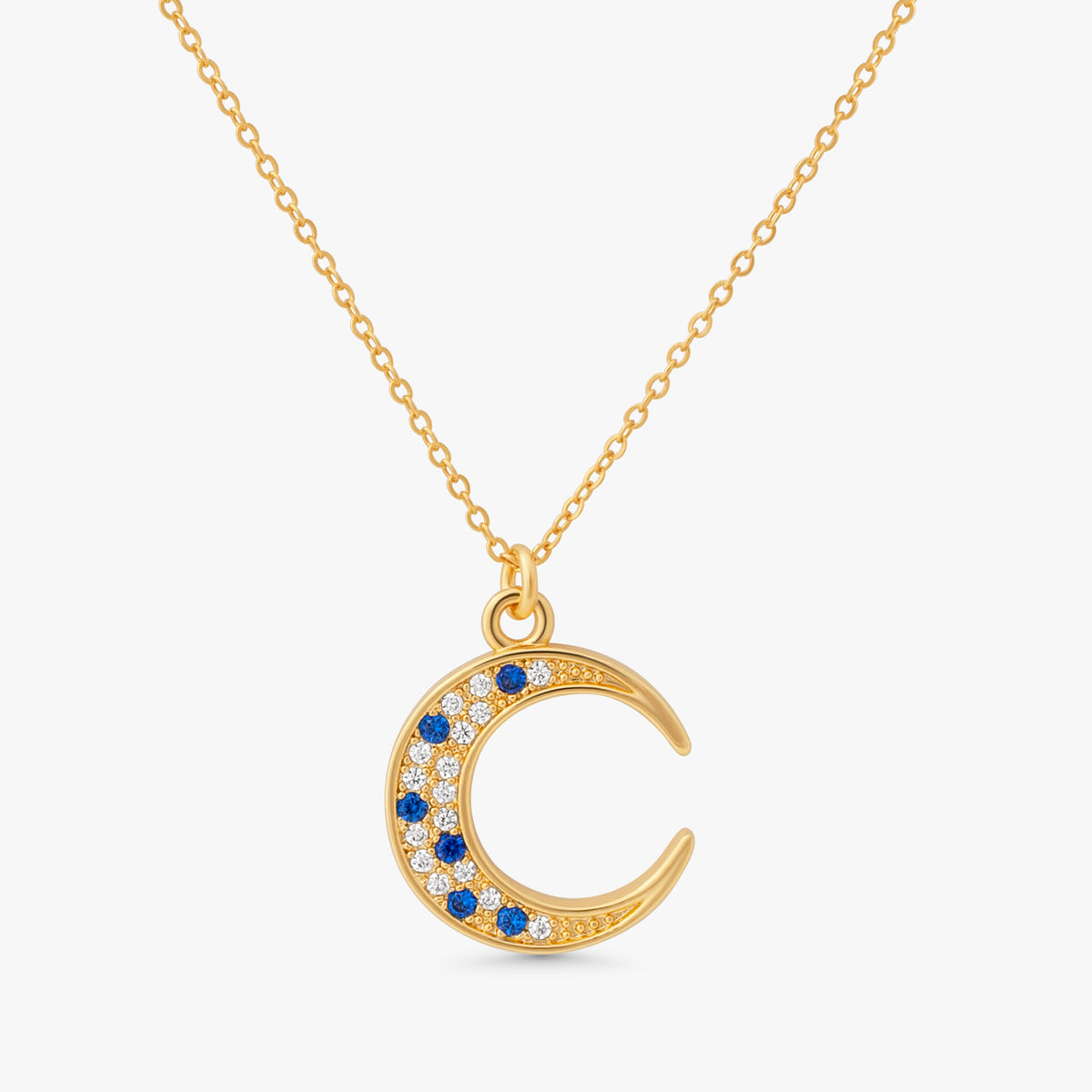 A gold crescent moon pendant necklace on a delicate gold chain, featuring small blue and clear gemstones along the curve, set against a light gray background.