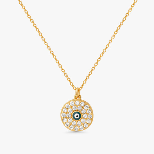 A gold chain necklace with a circular evil eye pendant, featuring clear pavé stones and a small green and white eye detail in the center, set against a light gray background.
