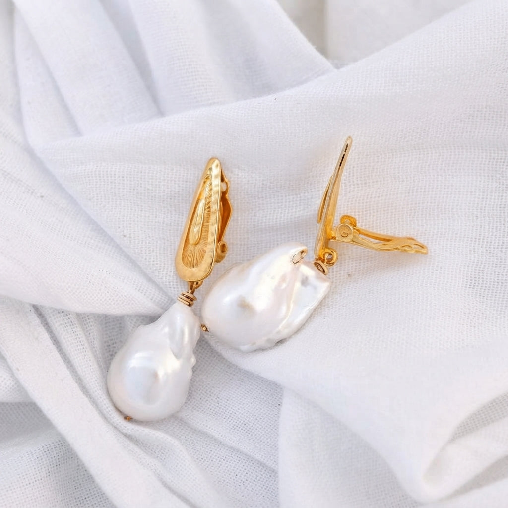 A pair of gold clip-on earrings with irregular white baroque pearl drops, displayed on a soft, draped white fabric background.
