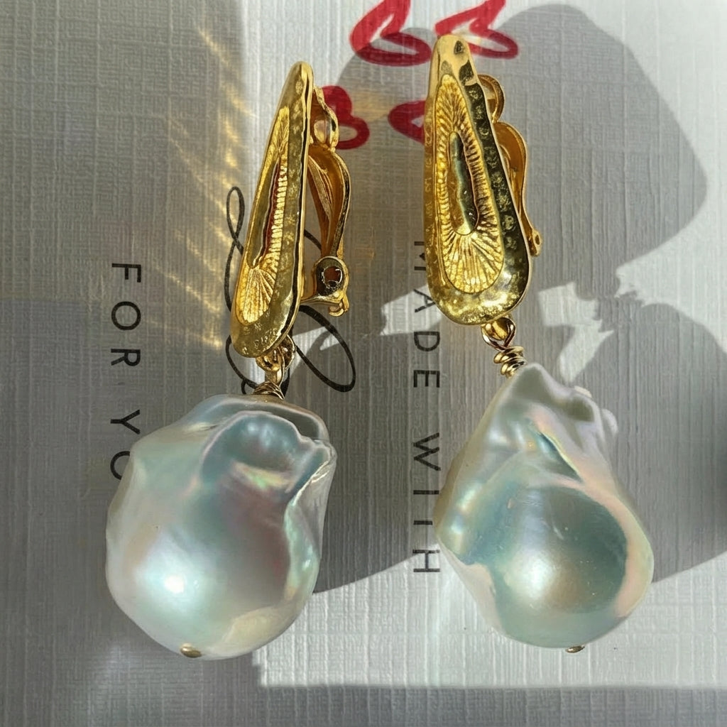 A pair of gold-toned clip-on earrings with large, irregular, pearly baroque pearl drops, displayed on a textured white surface with red heart graphics and shadows.