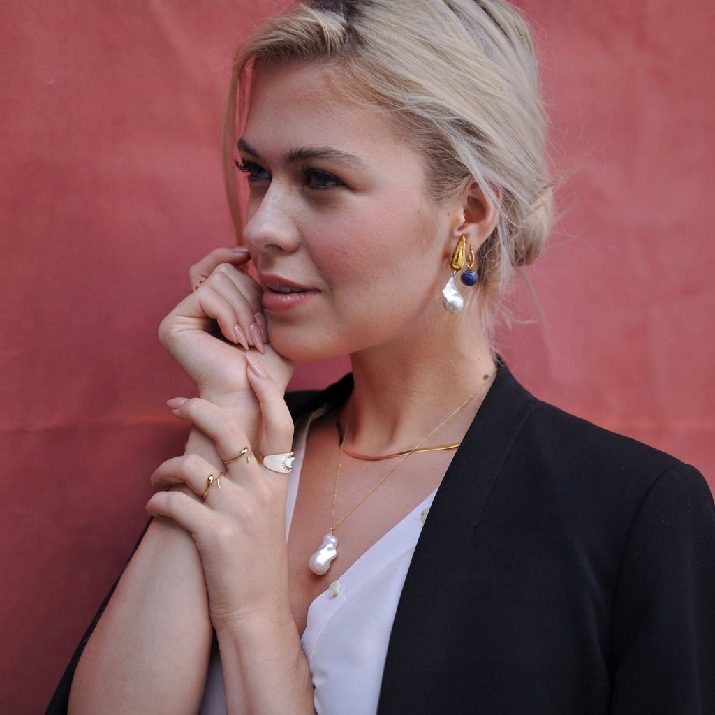 A blonde woman wearing gold and white jewelry, including a pearl drop earring, a gold necklace with a white pendant, and rings, posing against a textured reddish-pink background.