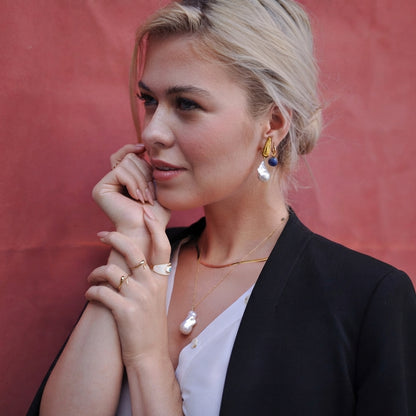A blonde woman wearing gold and white jewelry, including a pearl drop earring, a gold necklace with a white pendant, and rings, posing against a textured reddish-pink background.