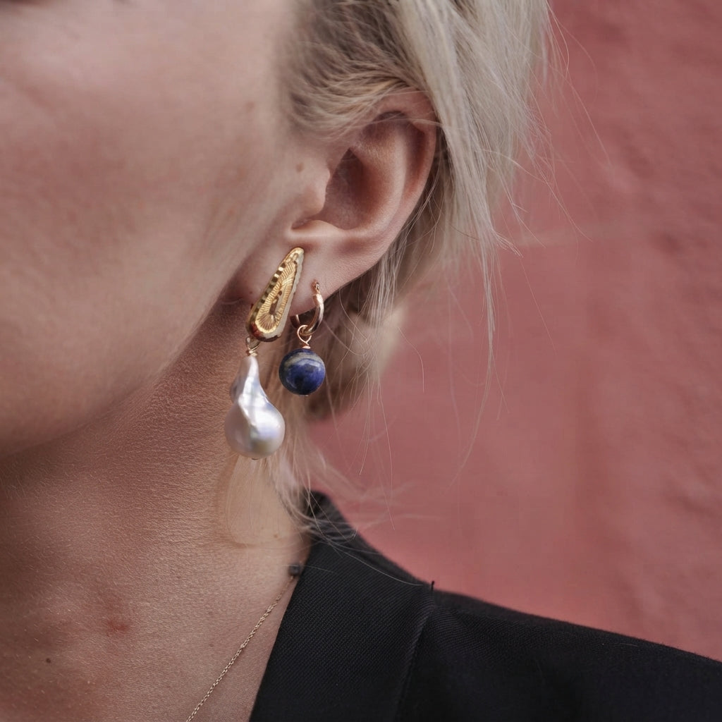 A close-up of a person's ear wearing a pair of gold huggie hoop earrings with a textured gold top piece, a white baroque pearl drop, and a small dark blue lapis lazuli sphere.