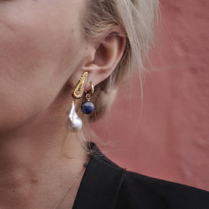 A close-up of a person's ear wearing a pair of gold huggie hoop earrings with a textured gold top piece, a white baroque pearl drop, and a small dark blue lapis lazuli sphere.