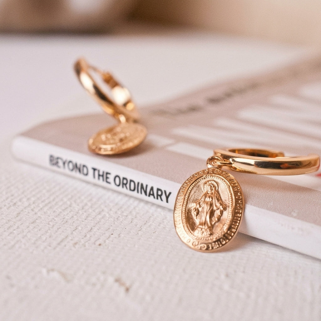 A pair of gold-filled huggie hoop earrings with oval religious medallions, displayed on a textured white surface next to a book titled 'BEYOND THE ORDINARY'.