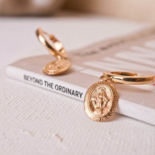 A pair of gold-filled huggie hoop earrings with oval religious medallions, displayed on a textured white surface next to a book titled 'BEYOND THE ORDINARY'.