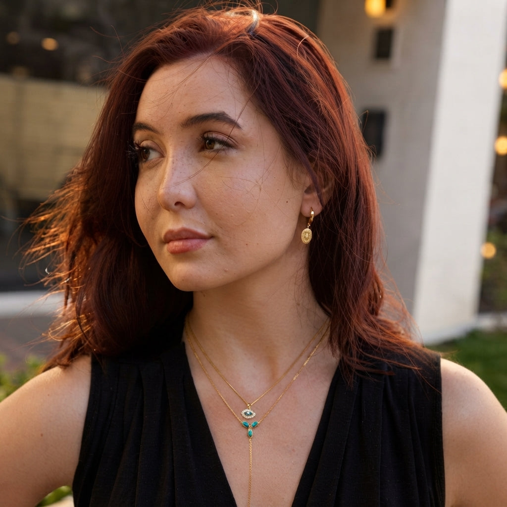 A woman with reddish-brown hair wearing a black sleeveless top and gold necklaces, including one with a turquoise and white evil eye pendant, photographed outdoors with a blurred background.