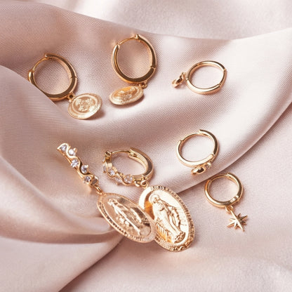 A set of gold-toned hoop earrings with various charms, including religious medallions, a star, and a rhinestone bar, arranged on a draped light pink satin fabric.