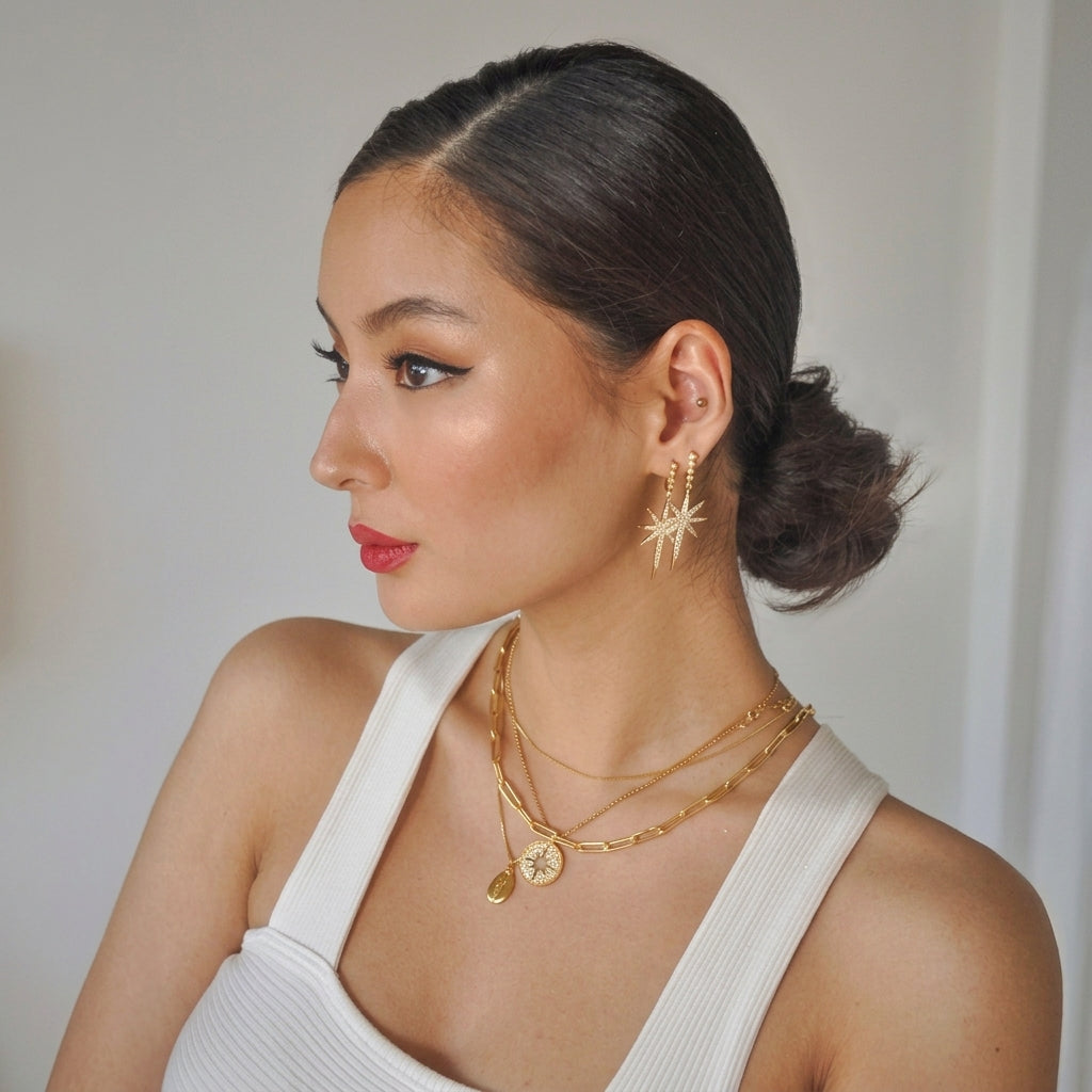 A woman with her hair in a low bun is shown in profile, wearing gold jewelry including a starburst earring, layered gold necklaces, and a gold ring, paired with a white ribbed tank top against a neutral light gray background.
