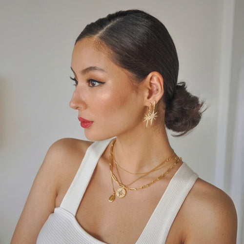 A woman with her hair in a low bun is shown in profile, wearing gold jewelry including a starburst earring, layered gold necklaces, and a gold ring, paired with a white ribbed tank top against a neutral light gray background.