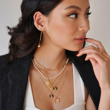 A close-up lifestyle shot of a woman wearing a layered gold necklace set with various charms, a gold dangle earring with a pearl, and gold rings, styled with a black blazer and white ribbed top.