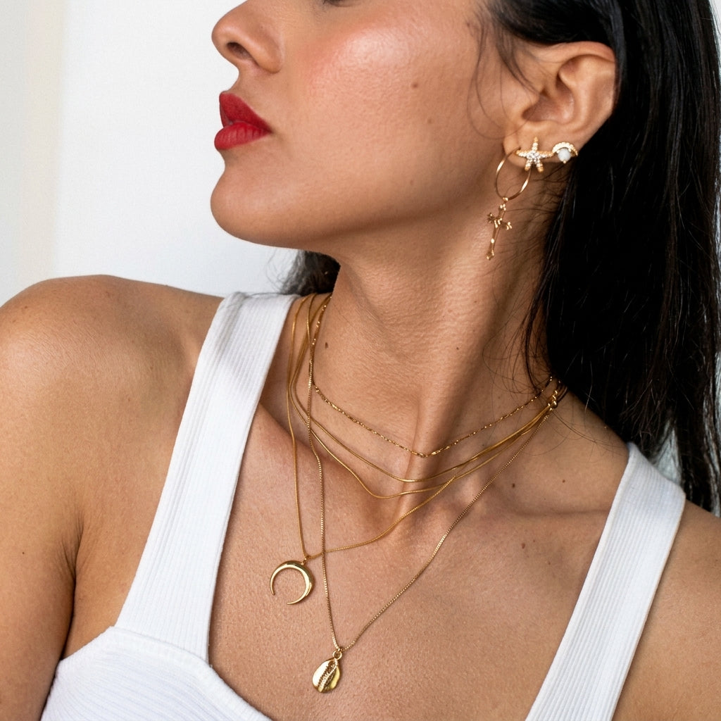 A close-up lifestyle shot of a woman wearing layered gold necklaces, including a crescent moon and teardrop pendant, along with matching gold hoop and star earrings.