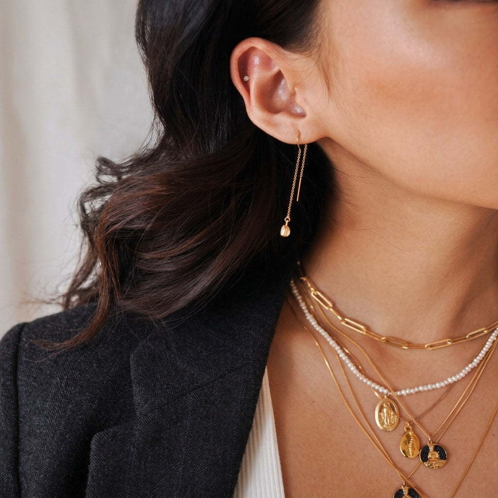A close-up lifestyle shot of a model wearing a delicate gold dangle earring, layered with gold and pearl necklaces, and dressed in a dark blazer.