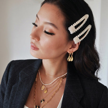 A woman wearing two pearl-bead hair clips in a side-swept hairstyle, styled with layered gold necklaces and gold drop earrings.