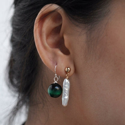 A close-up of a person's ear wearing two dangling earrings: one with a round dark green bead and one with a baroque-style white pearl, both attached to gold-toned hooks.