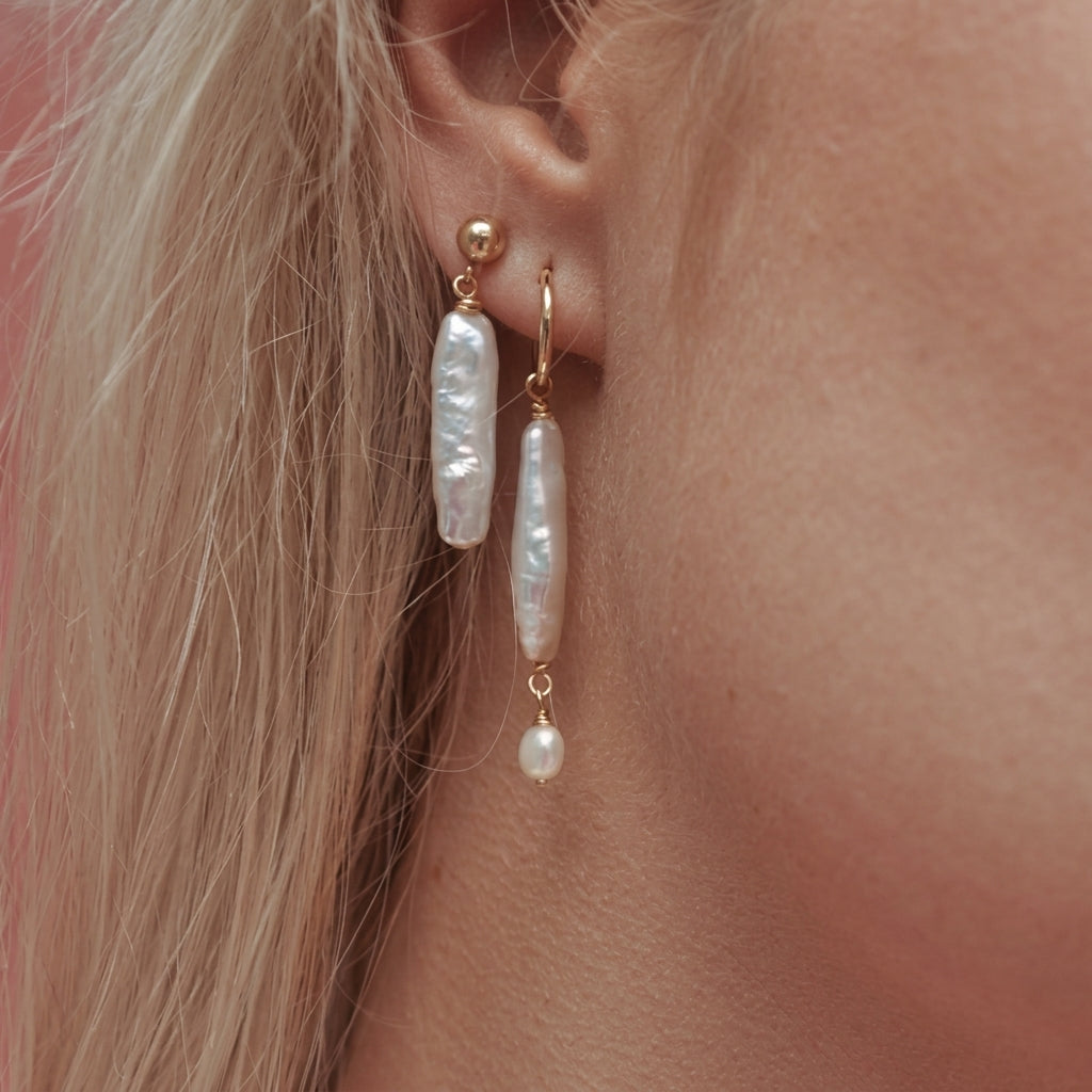 A close-up of a model's ear wearing two gold drop earrings with irregular white baroque pearls and small dangling pearl beads.