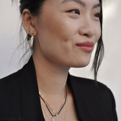 A close-up portrait of a woman wearing a gold huggie hoop earring with a textured, diamond-like silver and gold pavé design, along with layered gold necklaces, set against a plain light background.