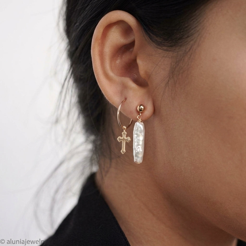 A close-up of a person's ear wearing a pair of gold hoop and drop earrings, including a small cross charm and a vertical bar with a textured white inlay, against a light background.