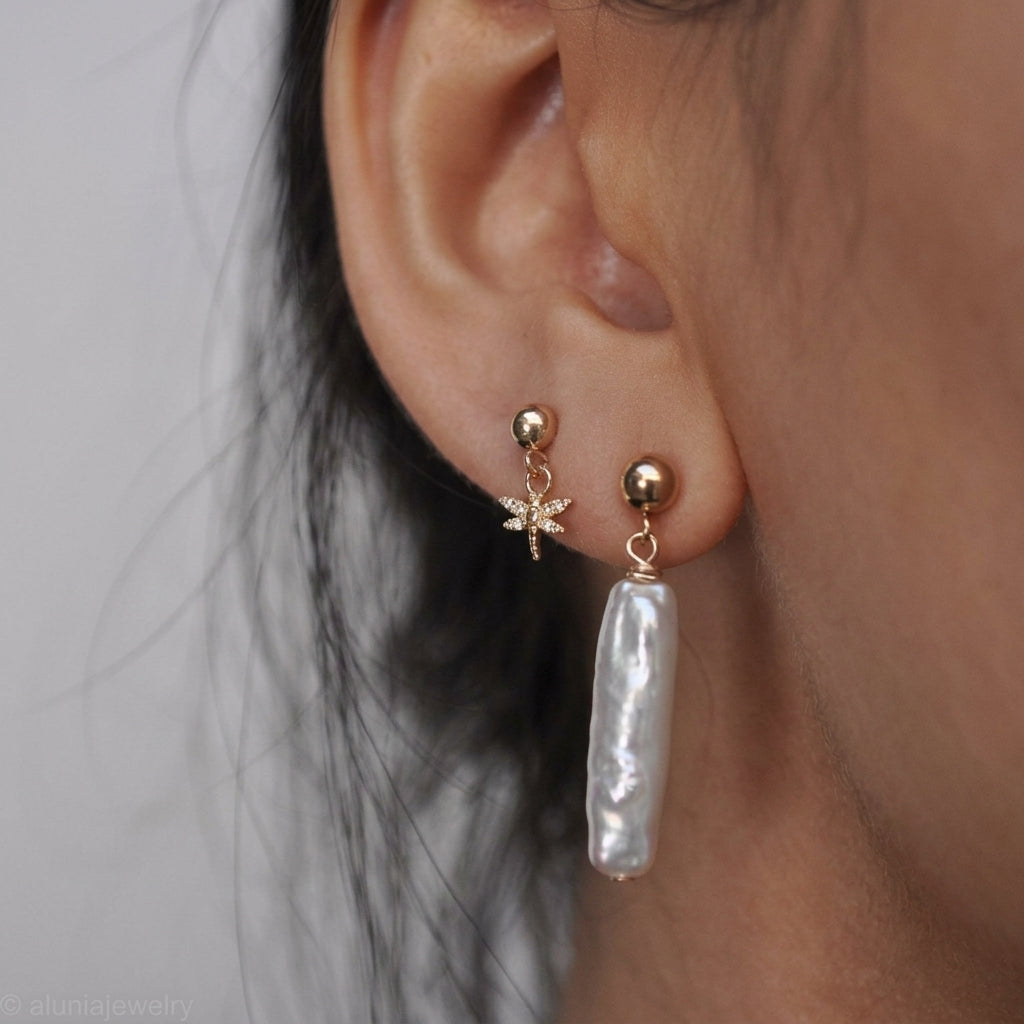A close-up of a person's ear wearing two gold-toned earrings: a small starburst stud earring and a dangling baroque pearl drop earring with a gold ring connector.
