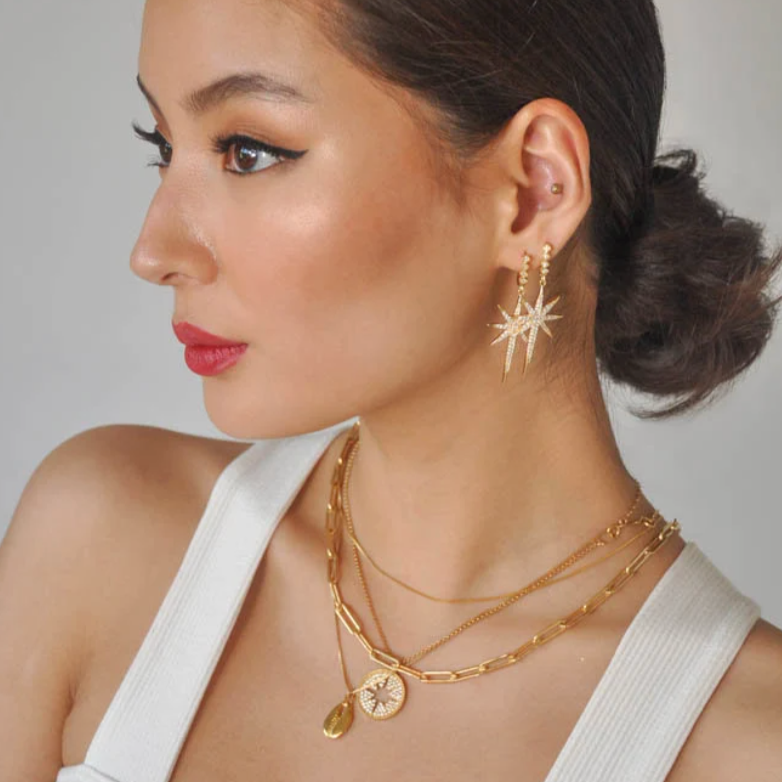 Model wearing gold starburst drop earrings paired with stacked gold chain necklaces.