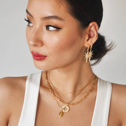 Model wearing white top with gold starburst earrings and layered gold chain necklaces.