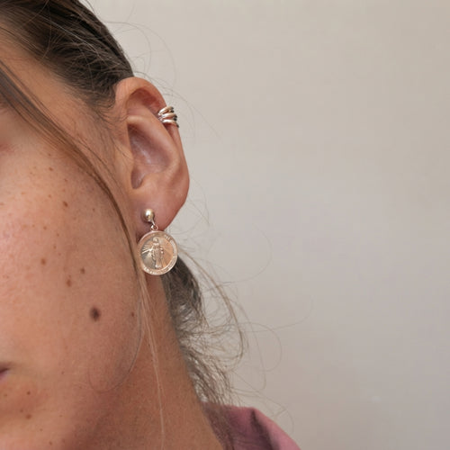 A close-up of a person's ear wearing a Gold -filled circular earring with a raised cross, alongside a stack of Gold ear cuffs, against a plain light gray background.