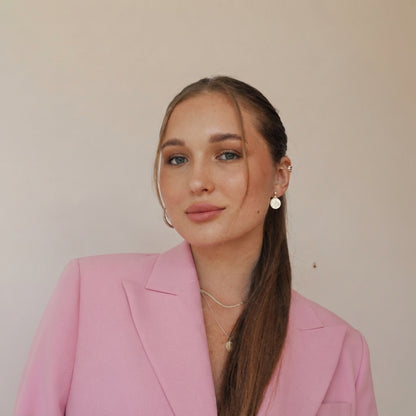 A woman with long brown hair in a ponytail wearing a pink blazer and delicate gold jewelry, posing against a plain beige wall.