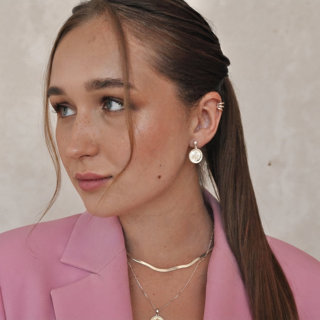 A woman with her hair in a ponytail wearing a gold circular drop earring, layered gold necklaces, and a gold ear cuff, paired with a pink blazer against a neutral beige background.