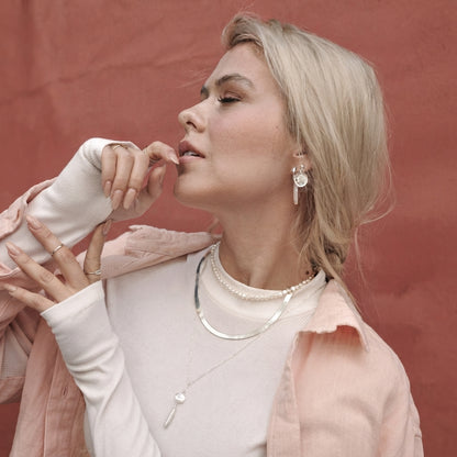 A blonde woman wearing layered silver necklaces and a dangling silver earring, posing against a textured terracotta background.