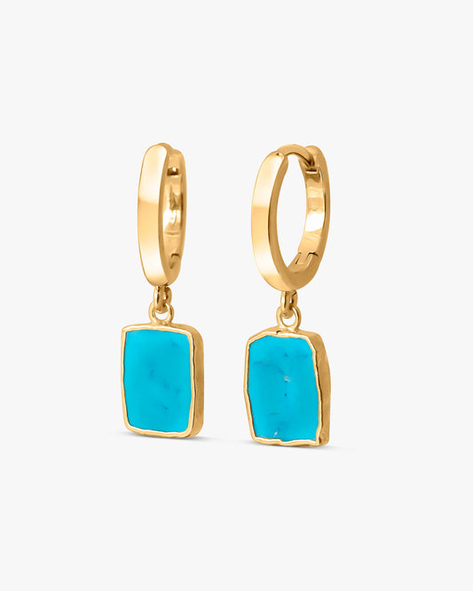 Pair of Gemma turquoise-tone huggie hoop earrings in 14k gold plating with rectangular dyed howlite charms, on a light gray background