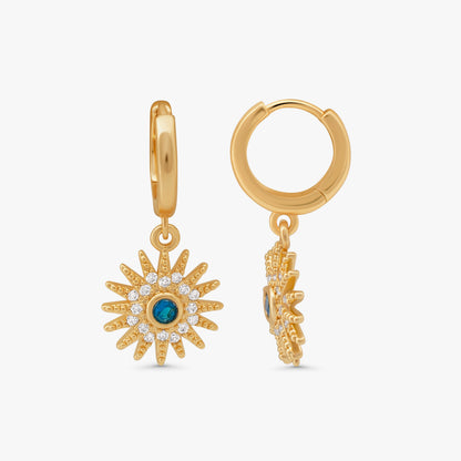 A pair of gold huggie hoop earrings with dangling sunburst charms. One earring is shown front-facing, featuring a gold sunburst design with clear pavé stones and a central round blue gemstone.